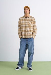 Rutet skjorte i gyldenbrun og creme, blå denim jeans og sort-hvide sneakers. Skjorten har en brystlomme og knapper.