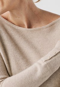 Maglione beige a maglia con scollatura off-the-shoulder e polsini a coste con dettagli di bottoni. Il materiale sembra morbido con un leggero luccichio.