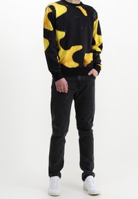 Sudadera negra con un patrón abstracto amarillo y negro, combinada con jeans oscuros y zapatillas blancas. Corte holgado, diseño de cuello redondo.