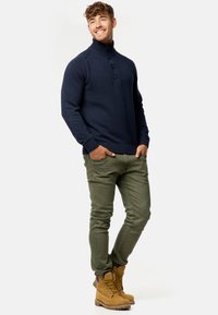 Mørkeblå striktrøje med høj krave og tre knapper, parret med olivengrønne slim-fit bukser og tan snørebåndsstøvler. Afslappet outfit.