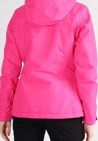 Světlý růžový voděodolný kabát s kapucí, nošený osobou z pohledu zezadu, doplněný o černé kalhoty.