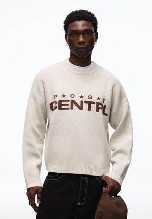 Giovane uomo che indossa un maglione crema con testo marrone "POST CENTRAL", pantaloni neri, tiene un berretto marrone in una mano su uno sfondo bianco semplice.
