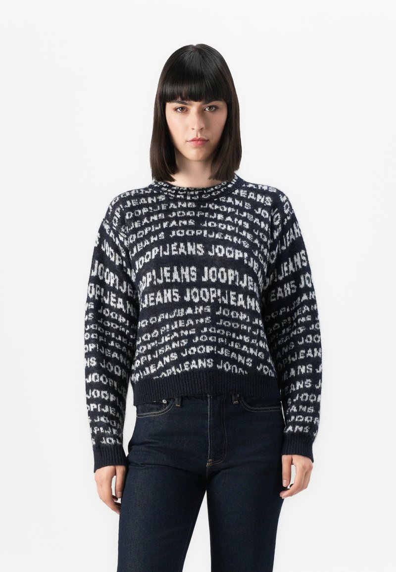 Ung kvinne med rett, mørkt hår iført en svart-hvit mønstret genser med teksten "JOOP! JEANS" og mørke tettsittende jeans.