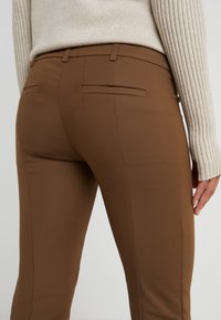 Close-up van een persoon die bruin aangesloten broeken en een beige geribbelde trui draagt, waarbij de achterzakken en riemlussen zichtbaar zijn.