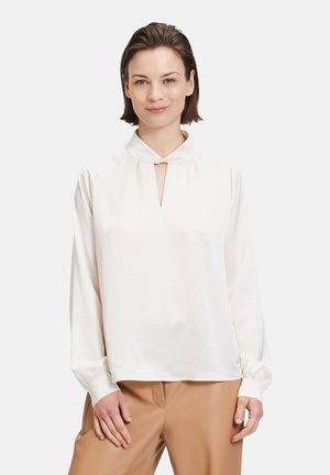 Weiße Bluse mit langen Ärmeln und einem Schlüssellochausschnitt, aus weichem Stoff, mit gerafften Schultern und Manschettenärmeln, getragen über beigen Hosen.