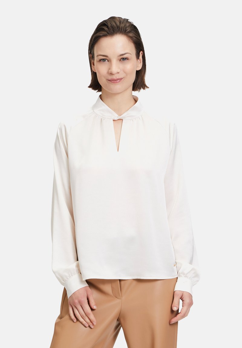 Blusa bianca a maniche lunghe con scollo a chiave, tessuto morbido, spalle arricciate e maniche con polsini, indossata sopra pantaloni beige.