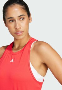 Top sportivo rosso in tessuto leggero, con scollatura rotonda e logo Adidas bianco. Indossato con un reggiseno bianco sotto.