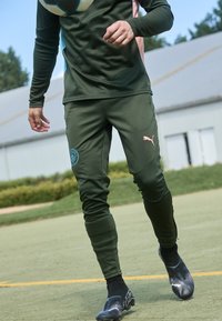 Tumši zaļi sporta bikses ar teksturētu audumu, ar zilu logotipa uzlīmi uz kreisā augšstilba un rozā akcentu uz labā gūžas. Melnie futbola apavi.
