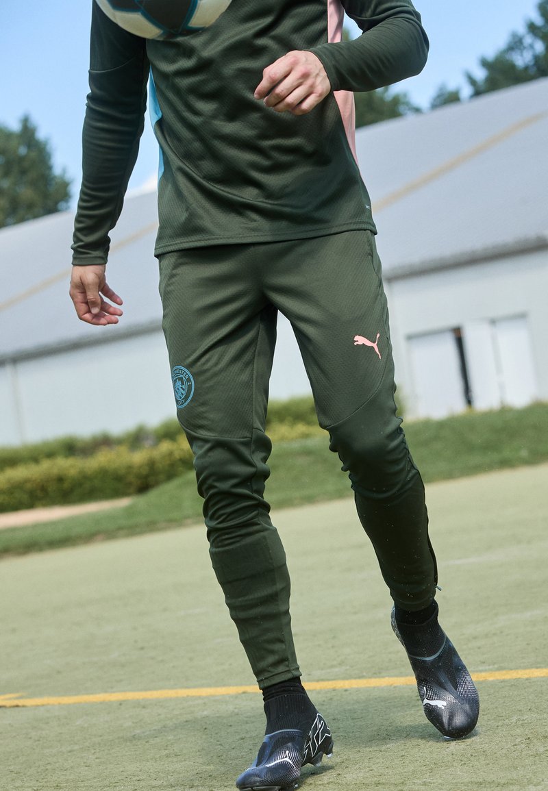 Tumši zaļi sporta bikses ar teksturētu audumu, ar zilu logotipa uzlīmi uz kreisā augšstilba un rozā akcentu uz labā gūžas. Melnie futbola apavi.