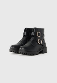 Black leather ankle boots with chunky soles, featuring two wide straps with gold buckles. Smooth texture and rugged tread pattern.