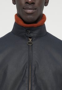 Ναυτικό μπουφάν με ψηλό γιακά και πορτοκαλί rib ύφασμα στον λαιμό. Διαθέτει φερμουάρ μπροστά με παλιού στυλ τράβηγμα και επίπεδη υφή υφάσματος.