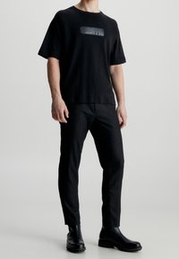 Svart t-shirt med kort ärm som har en glänsande logotyp. Kombinerad med mörka, slim-fit byxor och svarta ankelskor, vilket skapar en monokrom look.