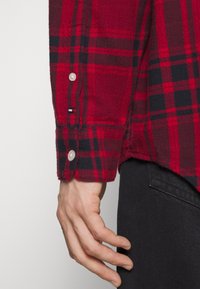 Chemise en flanelle à carreaux rouge et noir avec manches longues, poignets boutonnés et texture de tissu tissé. Présente une petite écusson logo près de l'ourlet.
