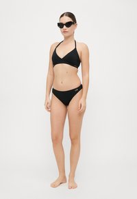 Melnā bikini komplekts ar trīsstūra formas halter neck topu un zemu vidukli. Gluda auduma ar zelta akcentu apakšā. Saulesbrilles noslēdz šo izskatu.