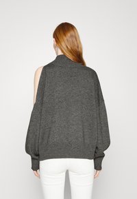 Nadměrný tmavě šedý svetr s vysokým stojáčkem, odkrytými rameny a žebrovanými manžetami, kombinovaný s bílými přiléhavými džíny. Jemná textura.