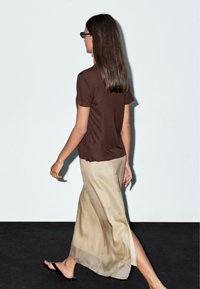 Brown short-sleeve top paired with a beige silky maxi skirt featuring a side slit. Model wears black flip-flops and sleek sunglasses.