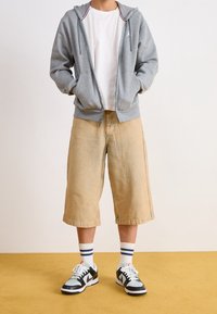 Felpa grigia con zip, t-shirt bianca, pantaloni corti beige fino al ginocchio, abbinati a sneaker bianco e nero e calzini a righe. Tessuti strutturati visibili.