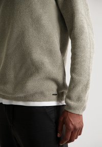 Hellbeige Strickpullover mit einem strukturierten Muster, gerippten Bündchen und einer weißen inneren Lage, die am Saum sichtbar ist. Schwarzes Etikett an der Seite.