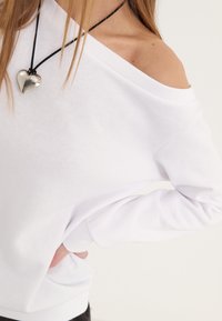 Sudadera blanca sin mangas con un diseño off-the-shoulder, con mangas largas y fabricada en un tejido suave. Presenta un corte relajado y puños acanalados. Incluye un collar con un colgante en forma de corazón plateado.