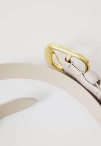 Vaalea beige nahkavyö, jossa on kultainen metallinen solki ja jonka pintarakenteessa on tekstuuria sekä kaareva muotoilu. Vyössä on "Calvin Klein" -brändäys.