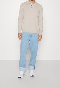 Beige långärmad polo med tre knappar, i kombination med ljusblå baggy jeans och vita sneakers, stående mot en neutral bakgrund.