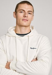 Felpa crema realizzata in morbido tessuto, con cappuccio regolabile, polsini a costine e un piccolo logo blu scuro "Pepe Jeans" sul petto.