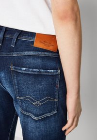 Dark blue denim jeans with back pockets featuring a zigzag stitch pattern, a leather tag with "REPLAY," and light wear on the fabric.