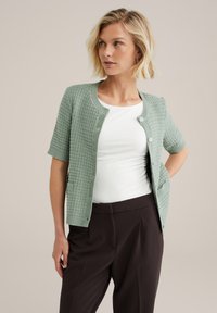 Gilet en tricot vert clair avec des manches courtes, motif texturé, encolure ronde et fermeture à boutons sur le devant. Porté sur un haut blanc et un pantalon sombre.