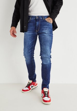 Man draagt strakke blauwe spijkerbroek met donkere vervaging op de kuiten, een zwart overhemd, wit T-shirt, rood-witte sneakers, en hand in de zak die een polstattoo toont.