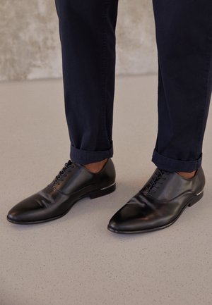 Zapatos de vestir de cuero negro con acabado liso, punta puntiaguda y diseño plano con cordones, combinados con pantalones azul marino doblados.