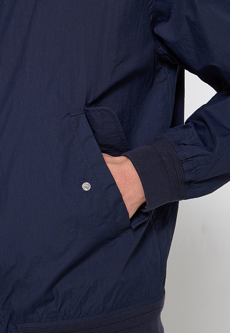 Dunkelblaue, leichte Jacke mit einem Reißverschluss vorne, gerippten Bündchen und seitlichen Taschen mit Druckknopfverschlüssen, gefertigt aus weichem, strukturiertem Stoff.