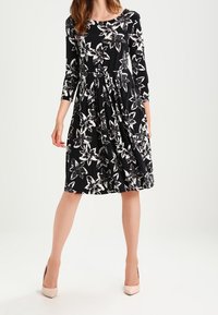 Robe fleurie noire à manches trois-quarts, taille cintrée et jupe mi-longue avec des motifs de fleurs blanches. Tissu à texture lisse.