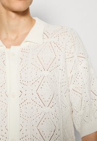 Gebreide polo-stijl trui in het wit met een gestructureerd ruitpatroon, met een knoopsluiting en korte mouwen.