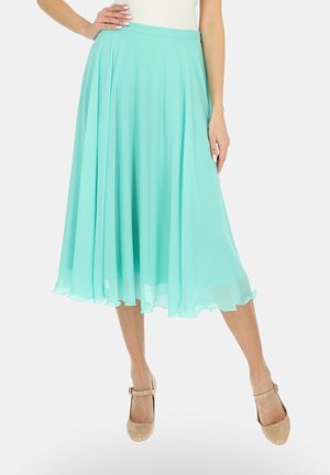 Kobieta w jasno-turkusowej spódnicy midi i beżowych czółenkach Mary Jane, stojąca z jedną ręką na biodrze.