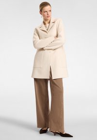 Cappotto di lana color crema con colletto a revers, due tasche frontali e design fino al ginocchio, abbinato a pantaloni larghi marroni.