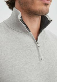 Sweat-shirt zippé gris avec col haut, intérieur gris foncé et fermeture éclair en métal. Le tissu a une apparence douce et texturée.