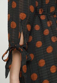 Černé šaty z látky s velkými rezavě zbarvenými puntíky. Obsahují nabíraný pas a zavázané manžety, s pomačkanou strukturou.