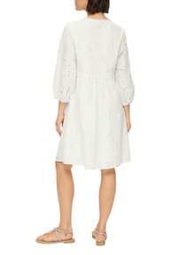 Robe en coton blanche avec un motif œillet en paisley, taille cintrée et manches bouffantes. Portée avec des sandales plates, montrant des jambes nues.