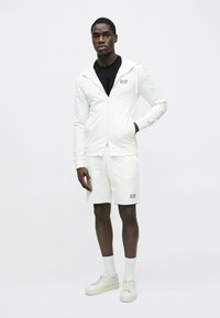 Bílá mikina na zip a odpovídající šortky z měkké látky. Výrazné logo EA7 na hrudi a šortkách, doplněné bílými teniskami a ponožkami.