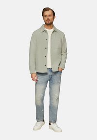 Hellgrüne Knopfjacke über einem cremefarbenen Hemd, kombiniert mit zerschlissenen blauen Jeans und weißen Sneakern. Verfügt über eine Brusttasche und Knöpfe.