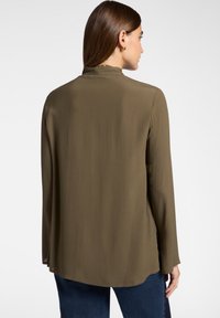 Donna con capelli castani lisci, che indossa orecchini a cerchio dorati, una blusa a maniche lunghe verde oliva e jeans blu scuro, che guarda verso il basso su uno sfondo bianco.