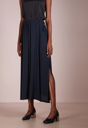 Vrouw draagt een zwart mouwloos topje, een marineblauwe midi-rok met een split aan de zijkant, en zwarte schoenen met blokhak, staand op een beige vloer.