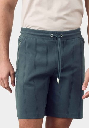 Mand iført mørkeblå shorts med snøre, lodret syning og sidelommer med lynlås, parret med en hvid skjorte.