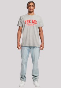 Gray cotton t-shirt with red lettering reading "THE MG CAR COMPANY," paired with light blue jeans and white sneakers, standing on a neutral background.