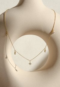 Złota naszyjnik z małymi charmsami, w tym kryształowym krzyżem, na delikatnych ogniwach, o gładkiej, zaokrąglonej sylwetce na teksturalnym tle.