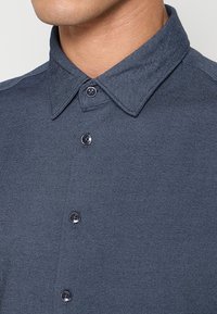 Donkerblauw overhemd met knopen, gemaakt van een gestructureerde stof, voorzien van een puntige kraag en vijf zwarte plastic knopen aan de voorkant.