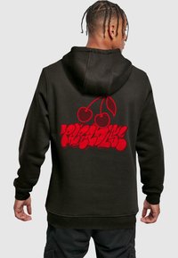 Osoba s copánky, oblečená v černé mikině s kapucí, na které jsou vzory červeného graffiti a grafika třešně na zádech, stojí čelem k nám.