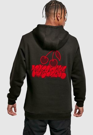 Person with braided hair wearing black hoodie with red graffiti-style text and cherry graphic on the back, standing facing away.