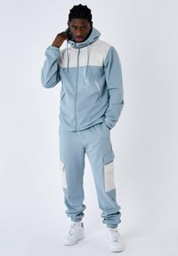 Felpa blu con pannello bianco, chiusura con zip sul davanti e cappuccio con lacci; abbinata a pantaloni jogging coordinati con tasche cargo beige.