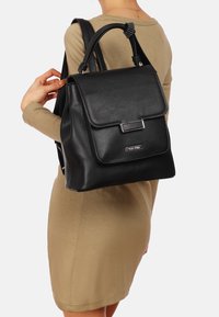 Zaino in pelle nera con chiusura a pattina, hardware argentato e dettagli testurizzati. Presenta spallacci regolabili e una forma strutturata.